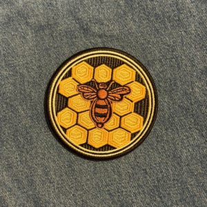 May include: A round embroidered patch featuring a honeybee design. The bee is orange and black, centered on a honeycomb pattern in shades of yellow and gold. The patch has a black border and is set against a denim background.