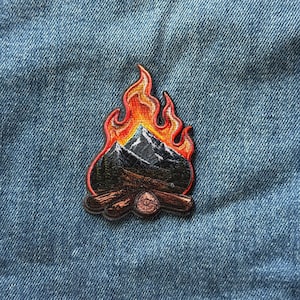 May include: An embroidered patch featuring a campfire scene with brown logs at the base, a dark mountain range in the center, and bright orange and yellow flames rising above the peaks, all set against a textured blue denim fabric background.