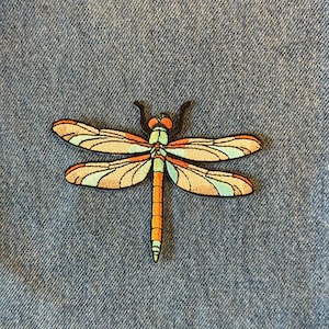 May include: A colorful dragonfly patch with orange, teal, and brown wings, and a segmented orange and green body. The patch is set against a blue denim background.
