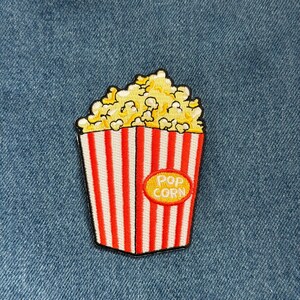 May include: Embroidered patch featuring a popcorn bucket with red and white stripes, filled with yellow popcorn. A yellow oval with the words "POP CORN" is on the front of the bucket. The patch is on a blue denim background.