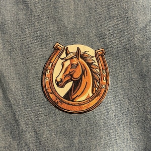 May include: Embroidered patch featuring a brown horse head within a horseshoe. The horse is set against a white background, with the horseshoe in a brown color. The patch is on a blue denim background.