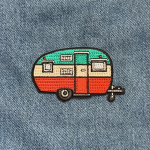 May include: Embroidered patch featuring a colorful camper design. The camper has a green roof, a cream-colored middle section, and a red bottom. The patch includes black outlines and a small wheel.