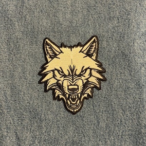 May include: Embroidered patch featuring a detailed wolf head with an open mouth, showcasing sharp teeth. The wolf's face is in beige with black outlines, set against a blue denim background. This patch is suitable for jackets, bags, or other fabric items.