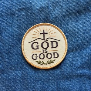 May include: A circular embroidered patch with a cream-colored background and a gold-colored border. The design features a dark brown cross above the text "GOD IS GOOD" in dark brown lettering, with a small green leaf sprig at the bottom.