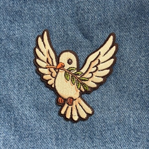 May include: Embroidered patch featuring a white dove with outstretched wings, holding an olive branch in its beak. The dove has brown outlines and details, set against a blue denim background. The patch is a symbol of peace.