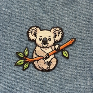 May include: Embroidered patch featuring a koala bear sitting on a brown branch with green leaves. The koala is gray and white with black accents, and the patch is set against a blue denim background.