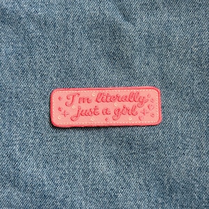 May include: A pink rectangular patch with the embroidered text "I'm literally just a girl" in cursive. The patch has a pink border and small embroidered stars and hearts. The patch is set against a blue denim background.