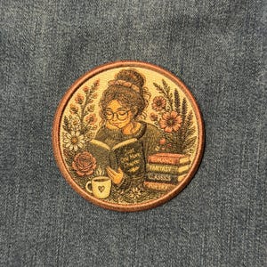 May include: A circular embroidered patch featuring a person with glasses reading a book titled Just One More Chapter, surrounded by flowers. A steaming mug with a heart and a stack of books labeled Romance, Fantasy, Classics, and Poetry are included.