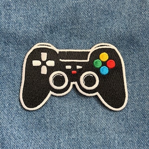May include: Black and white embroidered patch in the shape of a video game controller. The controller has a white outline, a directional pad, and colored buttons. The patch is on a blue denim background.