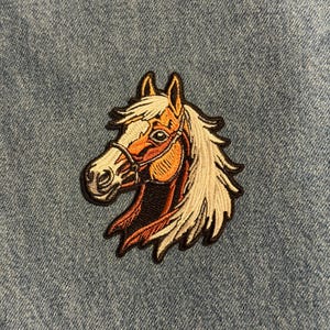 May include: Embroidered patch featuring a horse head with a tan and white mane, brown bridle, and detailed facial features. The patch has a black border and is set against a denim background.