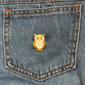May include: A gold-colored owl pin with white and yellow accents, set against a blue denim pocket. The pin features detailed feather patterns and bright yellow eyes. The pin is a decorative accessory.