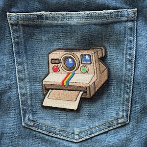 May include: An embroidered patch shaped like a vintage instant camera in shades of tan and brown, featuring a central blue lens, a red button, a green button, and a vertical rainbow stripe detail, attached to a blue denim fabric pocket.