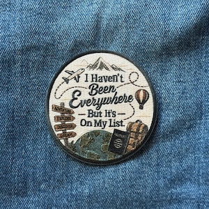 May include: A circular embroidered patch featuring a mountain peak, an airplane, a hot air balloon, a globe, a passport, and a backpack. The text reads: I Haven't Been Everywhere - But It's On My List. A signpost displays the words Adventure, Explore, Discover, Dream.
