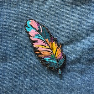 May include: An embroidered patch in the shape of a feather, featuring a vibrant color palette of pink, turquoise, yellow, orange, and purple threads. The feather design is outlined in black thread, creating a sharp contrast against the denim fabric.