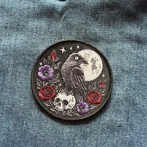 May include: A circular embroidered patch featuring a black raven perched on a white human skull. The design includes a large white full moon, small white stars, two red roses, one red rosebud, and two purple flowers against a black background.