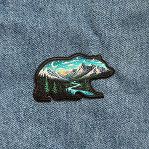 May include: Embroidered patch in the shape of a bear, featuring a mountain landscape with a river, trees, and a crescent moon. The patch has a black border and is set against a blue denim background.