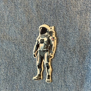 May include: An astronaut patch with a detailed design. The patch features a gray and white astronaut in a spacesuit, standing upright. The patch is set against a blue denim background.