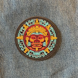 May include: Round embroidered patch featuring a stylized sun face design in red, yellow, and turquoise. The patch has a black border and is set against a blue denim background. The design is reminiscent of ancient art.