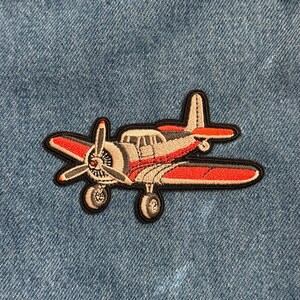 May include: Embroidered patch featuring a vintage airplane design. The patch has a red, white, and gray color scheme with black outlining. The airplane has a propeller, wings, and a tail. The patch is likely designed to be ironed or sewn onto fabric.