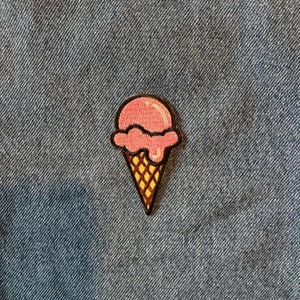 May include: Embroidered patch of a pink ice cream cone with a waffle cone base. The ice cream has a glossy finish and a dark outline. The cone is yellow with a brown outline. The patch is on a blue denim background.
