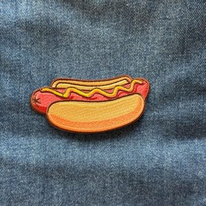May include: An embroidered patch shaped like a hot dog, featuring a red sausage, a tan bun, and a wavy line of yellow mustard. The patch is stitched with detailed thread work, showcasing distinct colors and textures for each food component.