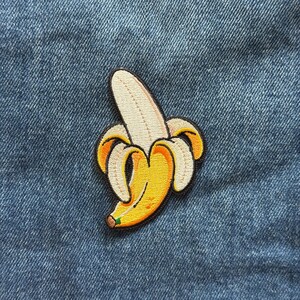 May include: An embroidered patch featuring a partially peeled banana with a bright yellow fruit body, a light cream-colored interior, and a dark brown stem tip. The design is outlined in black thread and is set against a textured blue denim fabric.