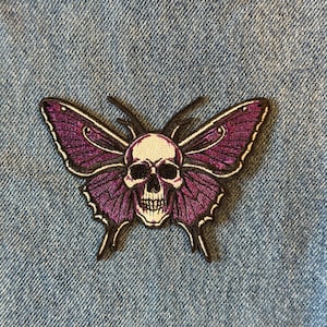 May include: Embroidered patch featuring a butterfly design with a skull in the center. The butterfly wings are purple with black outlines, and the skull is white. The patch is on a blue denim background.