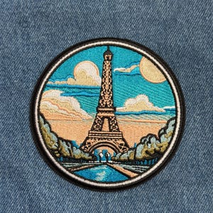 May include: Embroidered patch featuring the Eiffel Tower in Paris. The design includes a blue sky with clouds and a yellow sun, with the tower in the center. The patch has a black border and is set against a denim background.