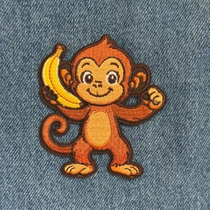 May include: Embroidered patch featuring a cartoon monkey holding a yellow banana. The monkey is brown and orange with a smiling face. The patch is on a blue denim background.