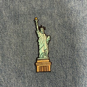 May include: Embroidered patch featuring the Statue of Liberty in shades of green, gold, and black. The patch depicts the iconic landmark with a torch held high, set against a denim background. A decorative item.