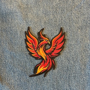 May include: Embroidered patch featuring a phoenix design. The bird is depicted with red, orange, and yellow colors, with black outlining. The patch is likely for clothing or accessories.