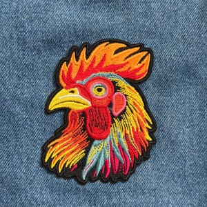 May include: Embroidered patch featuring a colorful rooster head. The patch has a black border and is set against a blue denim background. The rooster's comb and wattle are red, with yellow, orange, and blue accents.