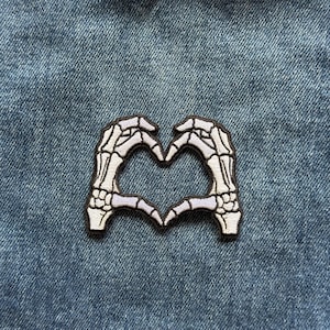 May include: An embroidered patch featuring two skeletal hands in a light gray and white color palette, positioned to form a heart shape with dark brown stitching outlining the edges of the bones against a blue denim fabric background.