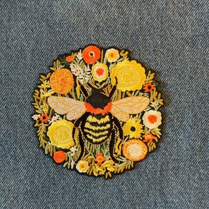 May include: Embroidered patch featuring a detailed bee surrounded by a wreath of yellow, orange, and white flowers. The bee has black and yellow stripes, with gray wings. The patch is on a blue denim background.