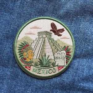 May include: A circular embroidered patch featuring a pyramid, a profile of a person in a feathered headdress, a stone carving, and a flying eagle. The design includes green, brown, and cream colors with the word MEXICO printed at the bottom.