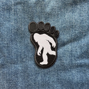 May include: An embroidered patch in the shape of a footprint with a black border and black toe sections. The center of the footprint features a white silhouette of a creature walking, set against a black background, attached to blue denim fabric.