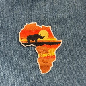 May include: Embroidered patch in the shape of the African continent. The patch features a sunset scene with a rhino silhouette, a large sun, and savanna animals. The patch is bordered with white stitching.
