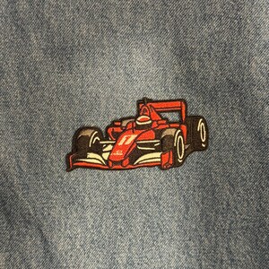 May include: A red and black embroidered patch of a Formula 1 race car with the number 17 on the side. The patch is on a blue denim background. The car has black tires and a white stripe.