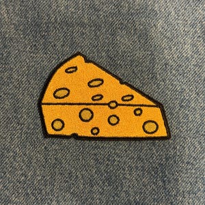 May include: Embroidered patch featuring a wedge of cheese with a yellow body and black outline. The cheese has several oval-shaped holes. The patch is set against a blue denim background.