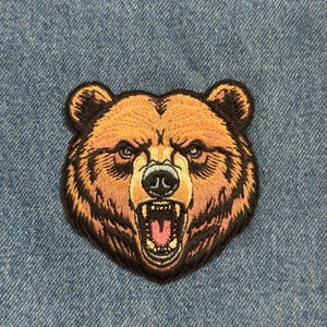 May include: Embroidered patch featuring a detailed bear head with an open mouth, showcasing teeth and a pink tongue. The bear has brown fur with black outlining and is set against a blue denim background. This patch is suitable for jackets, bags, or other fabric items.