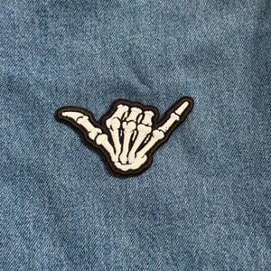 May include: A white skeleton hand patch with a black border, in the shaka gesture, is displayed on a blue denim background. The patch is an embroidered design, perfect for adding to clothing or accessories.