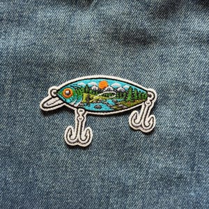May include: An embroidered patch shaped like a fishing lure with a white border. The design features a landscape with a blue river, green pine trees, snow-capped mountains, and an orange sun. Two white metal-style hooks hang from the bottom.