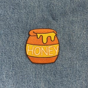 May include: An embroidered patch featuring a cartoon honey pot with dripping honey. The pot is orange with a yellow band that reads "HONEY" in white letters. The patch is set against a blue denim background.