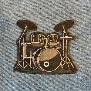 May include: A black and gray embroidered patch featuring a drum set. The patch shows a bass drum, snare drums, cymbals, and stands. The patch is on a blue denim background.