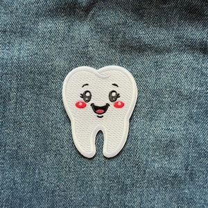 May include: A white embroidered patch in the shape of a tooth with a smiling face. The tooth features black embroidered eyes with eyelashes, a black mouth, and two small red circular cheeks, all set against a textured blue denim fabric background.