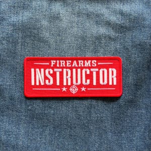 May include: A rectangular red embroidered patch with white stitched text that reads FIREARMS INSTRUCTOR. The design includes a horizontal line above and below the text, with two small stars and a central target symbol at the bottom.