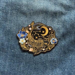 May include: An embroidered patch featuring a gold koi fish swimming amidst swirling water, a crescent moon, and small stars on a black background. The design is accented with blue and light blue flowers and small pink blossoms, all outlined in black.