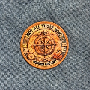 May include: Round patch with a compass design and the text "NOT ALL THOSE WHO WANDER ARE LOST." The patch features a compass rose, a map, and a mountain illustration. The patch is brown and orange with a stitched border.