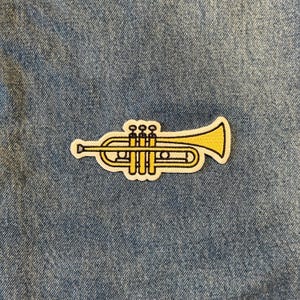 May include: A yellow trumpet patch with a white border is centered on a blue denim background. The patch features detailed lines and curves, showcasing the instrument's design. The patch is likely an iron-on or sew-on accessory.