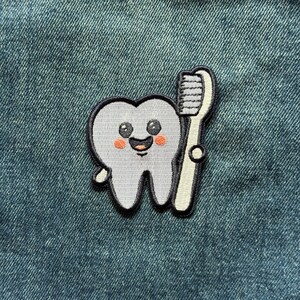 May include: An embroidered patch featuring a light gray cartoon tooth with a smiling face, black eyes, and pink cheeks, positioned next to a white toothbrush with gray bristles. The patch has a thick black border and is stitched onto blue denim fabric.